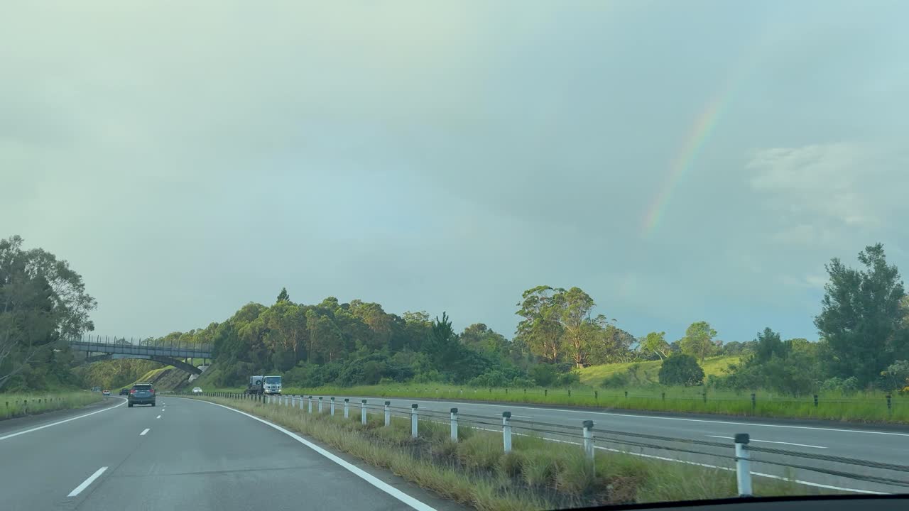 A vehicle travels down a multi-lane rural highway under overcast skies, with a faint rainbow visible above green fields and scattered trees. Steady forward camera movement