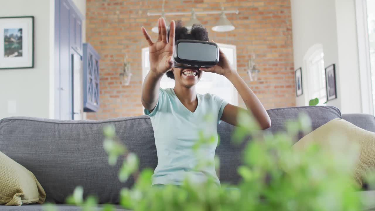 Happy african american teenage girl sitting on sofa, wearing vr headset and touching virtual screen