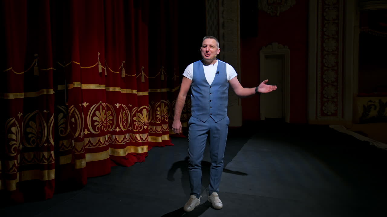 Man in front of empty theatre hall with red velvet chairs. Actor is standing on stage and talking with gestures before the performance.