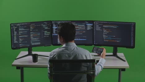 vista trasera de un desarrollador asiático trabajando con un teléfono móvil mientras escribe código con múltiples pantallas de computadora en el estudio de pantalla verde