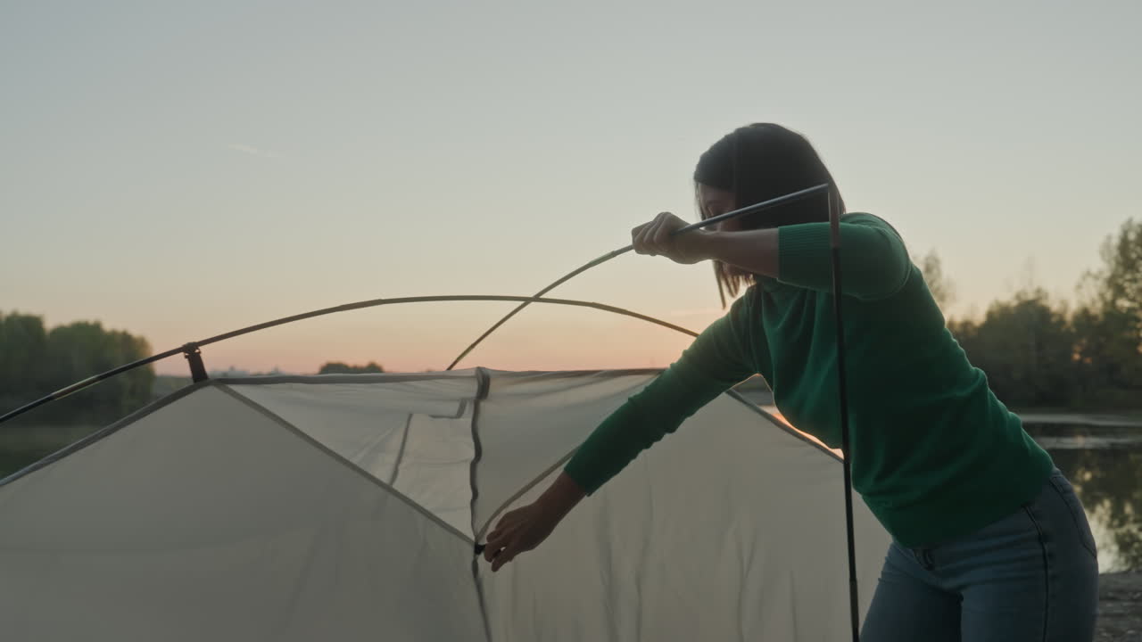 Campingtelt samlet af kvinde ved søbredden i solnedgang, hænder fører stænger igennem og sikrer stof på stenet bred, fokuseret camper forbereder aftenens lejrplads, blødt gyldent lys over den rolige sø