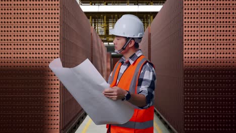 side view of asian male engineer with safety helmet looking at blueprint in his hands and looking around while standing with red brick packed in stacks are stored