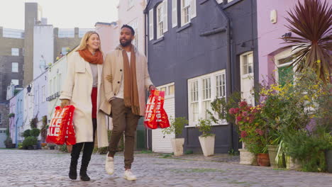 Multi-cultural-couple-arm-in-arm-as-they-walk-along-cobbled-mews-street-on-visit-to-city-in-autumn-or-winter-carrying-sale-shopping-bags---shot-in-slow-motion