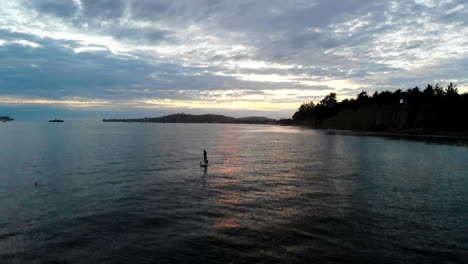 luftdrohnenaufnahme bei sonnenuntergang, tief über den bunten spiegelungen des wassers des pazifischen ozeans fliegend, mit einem paddelboarder in silhouette entlang der kalifornischen strandklippen von santa barbara, kalifornien