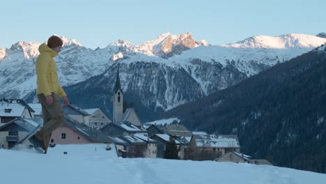 young caucasian man in yellow jacket and beanie walking and stopping to see view of small mountain village and church in guarda, switzerland