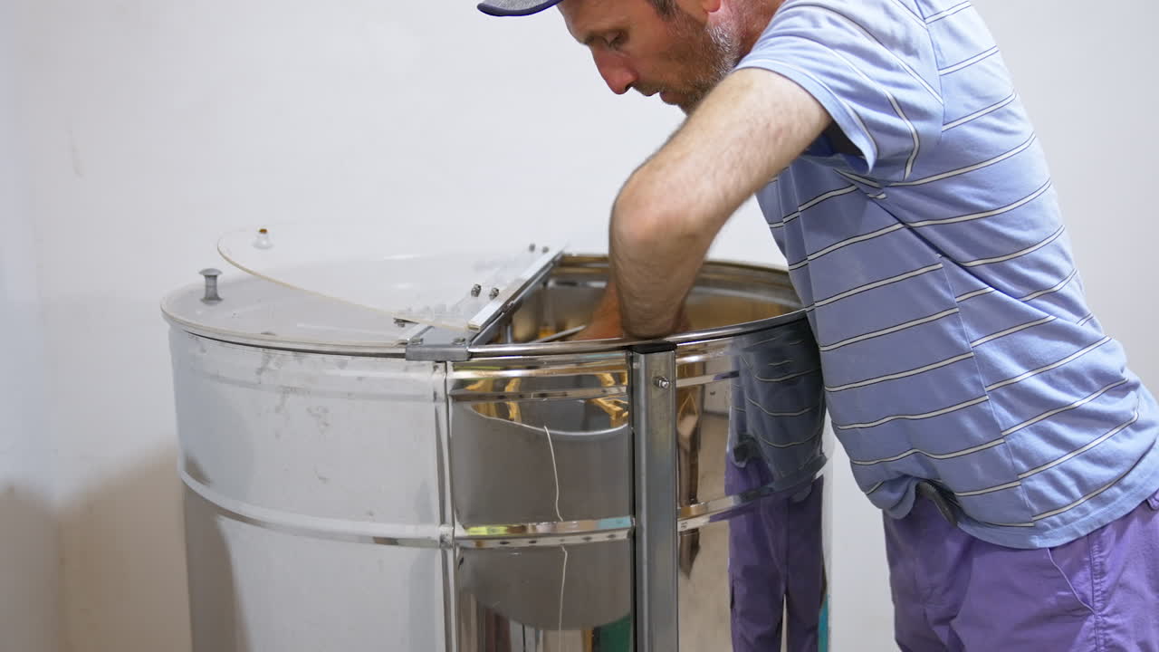 Apiarist preparing frames for honey extracting form cells. Male farmer places frames in a big metal barrel-like apparatus.