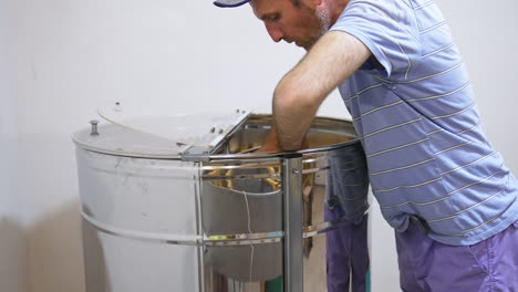apiarist preparing frames for honey extracting form cells. male farmer places frames in a big metal barrel-like apparatus.