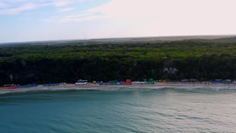 Magnífica-Y-Amplia-Plataforma-Aérea-Para-Drones-Y-Toma-Inclinada-Hacia-Arriba-De-La-Playa-Tropical-Praia-Do-Madeiro-Con-Coloridas-Sombrillas-Cerca-De-La-Famosa-Ciudad-De-Pipa-En-El-Norte-De-Brasil