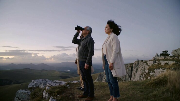 Scanning man in sweater raising binoculars surveying hills at cliff as dusk falls, cardigan moving