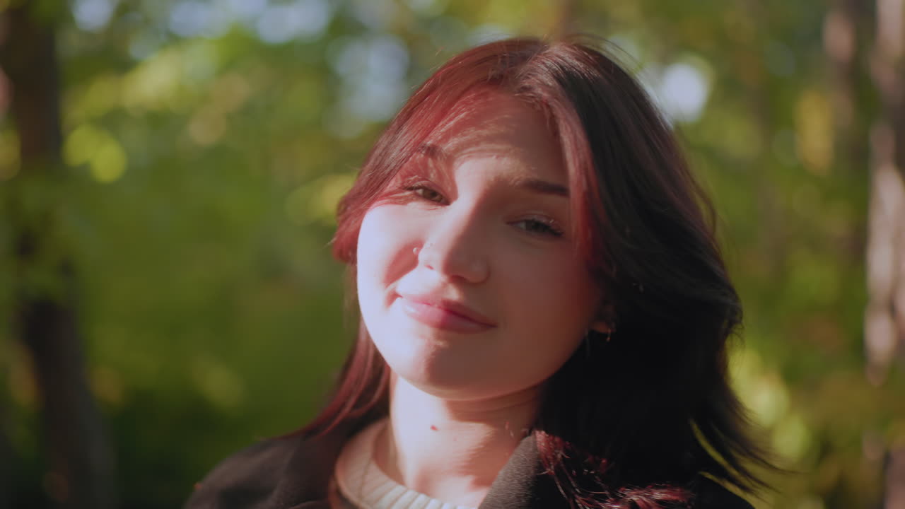 Cheerful woman tilts head in relief under warm sunlight, eyes closed, soft smile bright on face, forest bokeh around, calm moment of gratitude captured in serene autumn setting