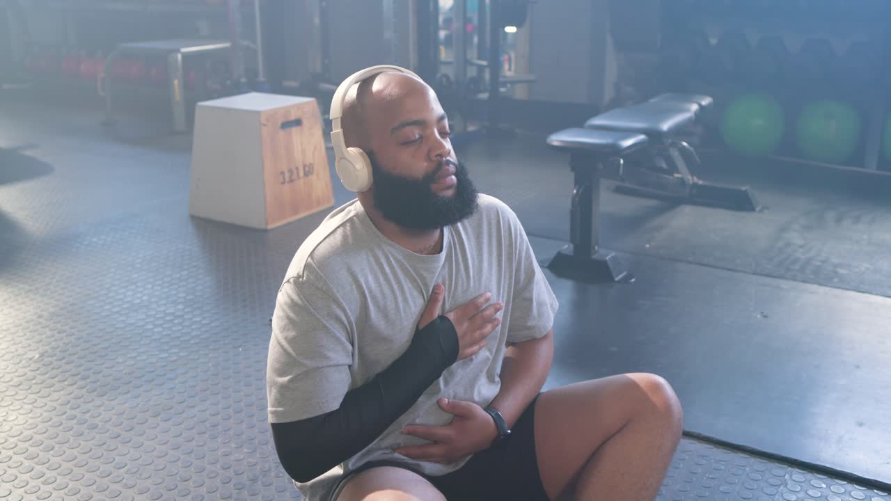 Pria keturunan Afrika-Amerika meletakkan tangan di dada dan perutnya sambil melakukan kontrol pernapasan di gym, mengenakan headset.