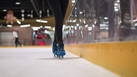 close view of a woman wearing black trousers and blue skates, turning slowly on an ice rink, she has black nail polish on her right hand, other skaters with blurred light reflections around