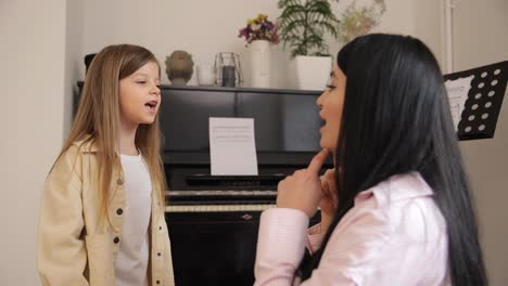 a woman is instructing a young girl in singing while sitting at a piano. in the room, a woman is joyfully teaching a little girl how to sing while they stand next to a piano together