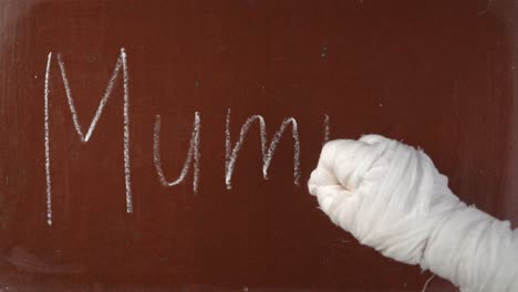 textured brown chalkboard background mummy's hand in bandages writing 'mummy' on the board with a