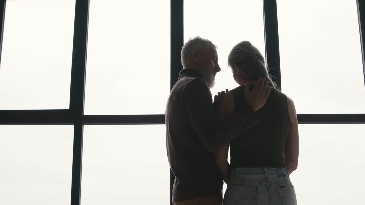 Back View Of Middle Aged Man Embracing His Sad Wife And Trying To Comfort Her While Standing Together At Window At Home 1