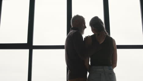 vista trasera de un hombre de mediana edad abrazando a su triste esposa y tratando de consolarla mientras están juntos en la ventana de casa 1