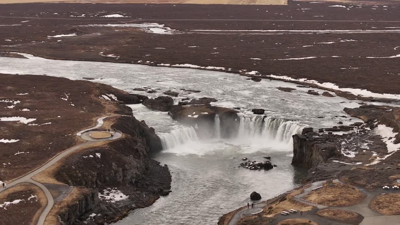 Sweeping aerial view of Goðafoss waterfall on the Skjálfandafljót River near Laugar, Iceland, with powerful water plunging into a wide canyon framed by early spring tundra.