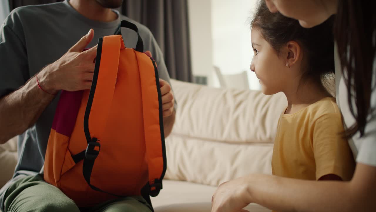 Close-up shot of a brunette man dad in a gray T-shirt putting a bright school backpack on his little daughter in a yellow dress. Happy mother of a girl hugs her, in a modern apartment