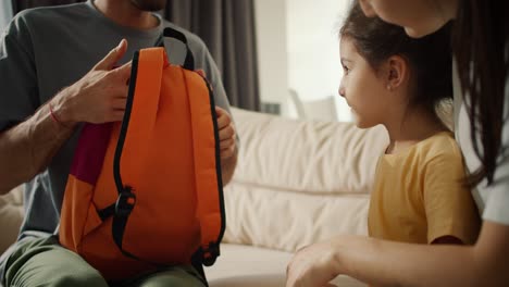 una toma de primer plano de un hombre moreno padre en una camiseta gris poniendo una mochila de la escuela brillante en su hija pequeña en un vestido amarillo. madre feliz de una niña la abraza, en un apartamento moderno