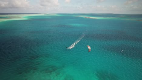 A-kite-surfer-on-vivid-blue-waters-with-a-boat-nearby,-sunny-day,-aerial-view