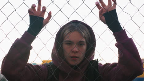 exercise lover wearing maroon hoodie and black vest gripping chain link fence with both hands during short rest after intense outdoor workout on urban sports court at dusk under city lights