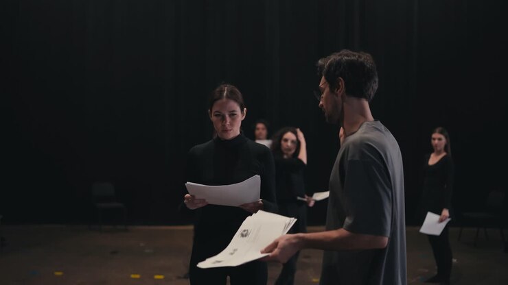 A confident young brunette man the stage director tells how to play his role correctly and say the text of a brunette girl to an actress in a black suit during general preparation and rehearsal for a performance in a theater on stage