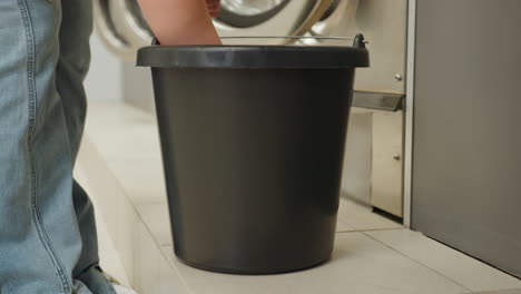 Lower view of laundry owner in jeans carrying bucket with blue towel, squeezing water before cleaning near industrial washing machines, service worker prepping for sanitation in modern laundromat