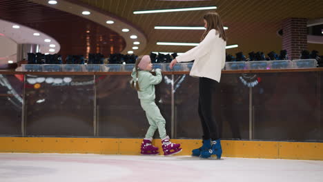 a close view of a young girl and her mom standing on an ice rink, the girl is dressed in a mint green outfit and pink skates, behind them is a row of rental skates and a blurred festive background