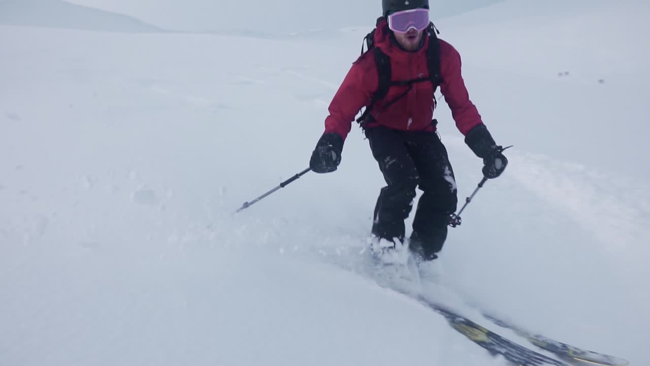 toma en cámara lenta de un hombre con chaqueta roja, esquiando cuesta abajo, rodeado por una montaña blanca como la nieve y un fondo semisoleado cubierto de nubes-1