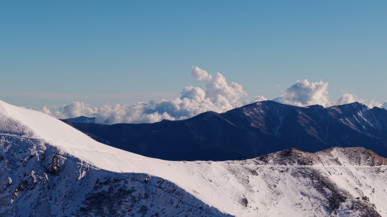 Drone terbang di atas Puncak Alpen yang Bersalju dan Medan Pegunungan Tinggi, langit siang hari yang biru