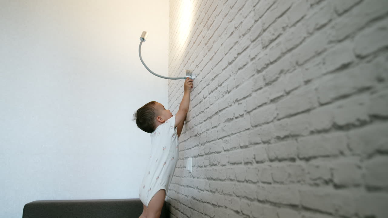 Caucasian toddler in white bodysuit turns on the lamp on the wall. Baby plays with light at home.