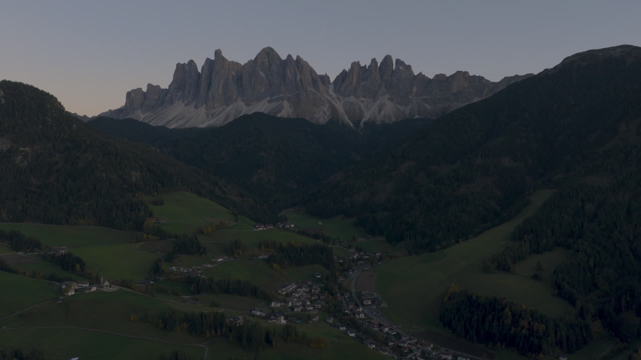 Vista aerea cinematografica delle Dolomiti italiane all'alba, aspre cime rocciose sopra rigogliose colline alpine e chalet sparsi, sereno paesaggio naturale di montagna