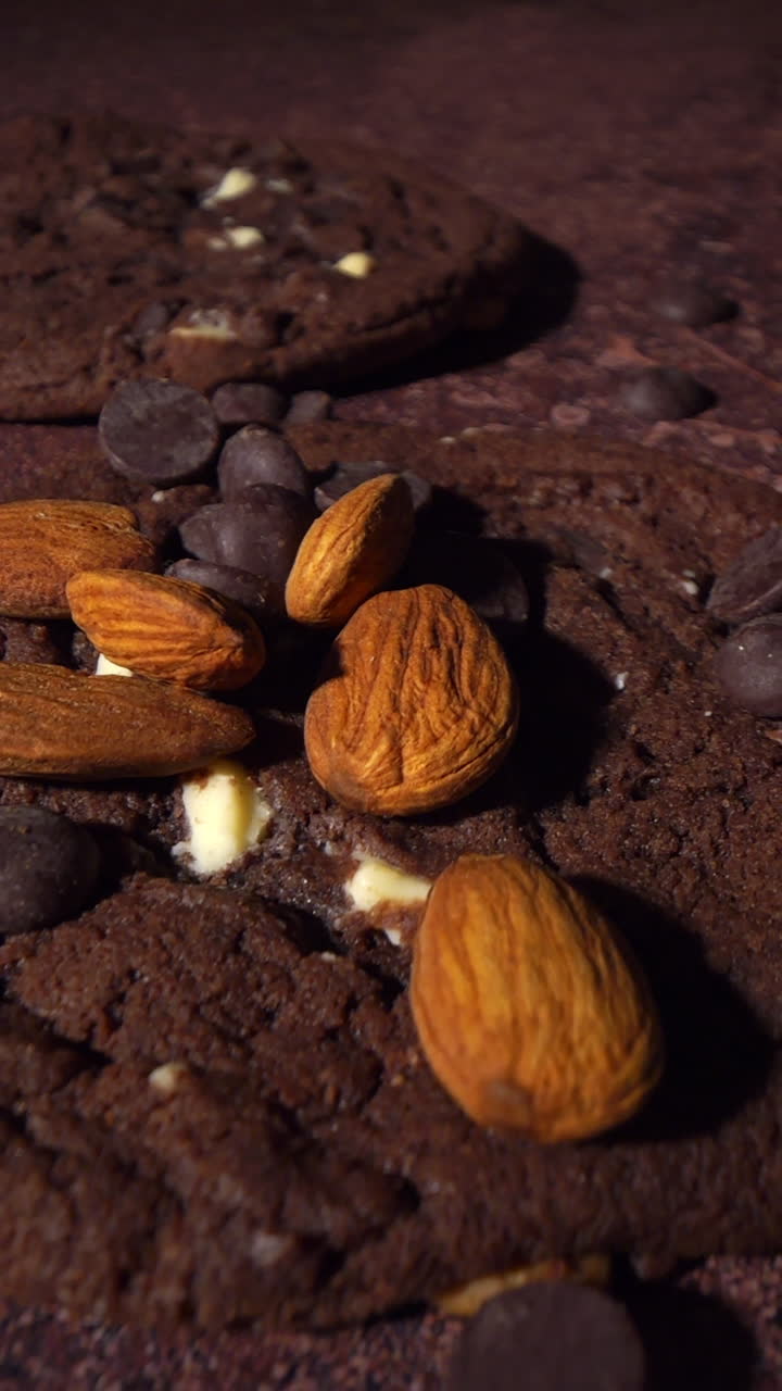 Kamerabevegelse i sakte film over sjokoladekjeks mens mandler og sjokoladebiter faller ned på den bakte overflaten. Detaljert makrovisning av smuler, tekstur og ingredienser i bevegelse.