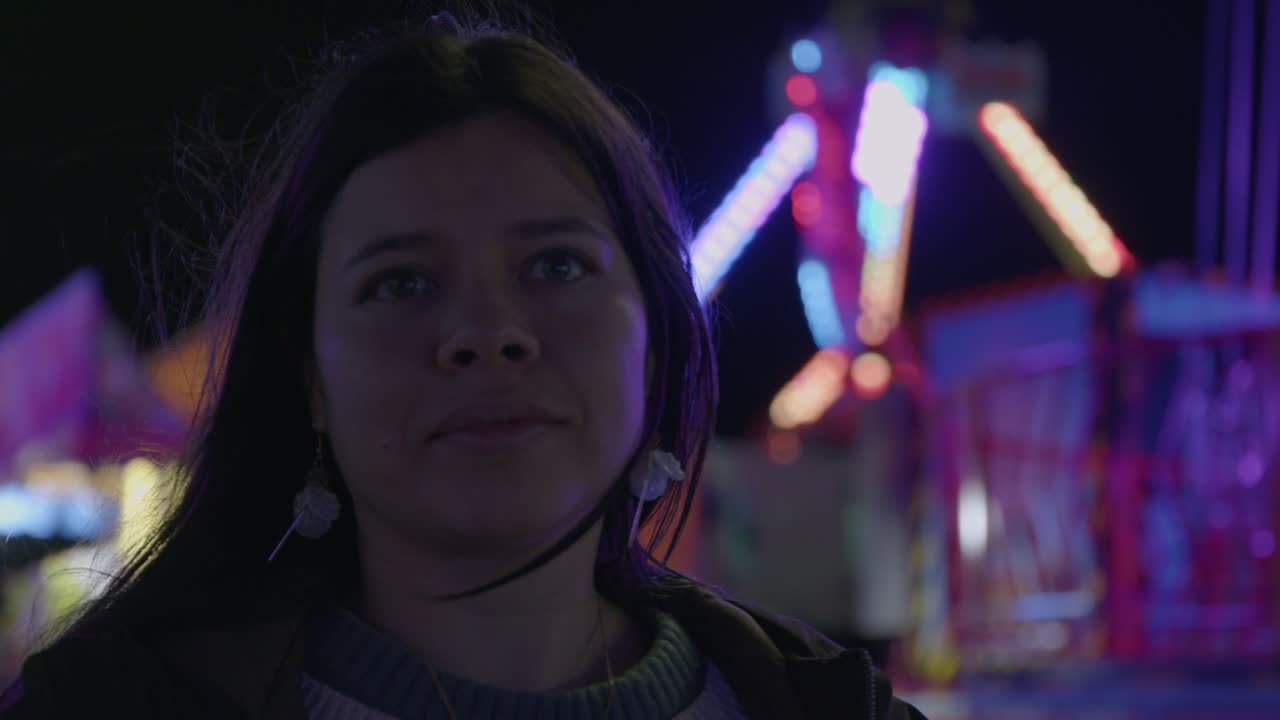 Uma bela foto de uma jovem aproveitando a atmosfera vibrante de um carnaval à noite. Ela observa os brinquedos coloridos e fora de foco com uma sensação de admiração e alegria.