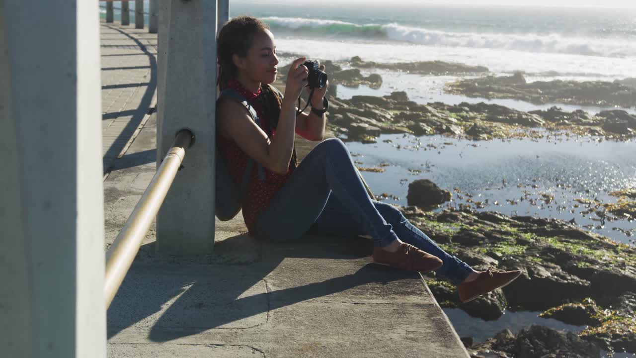 African american woman sitting and photographing on promenade by the sea