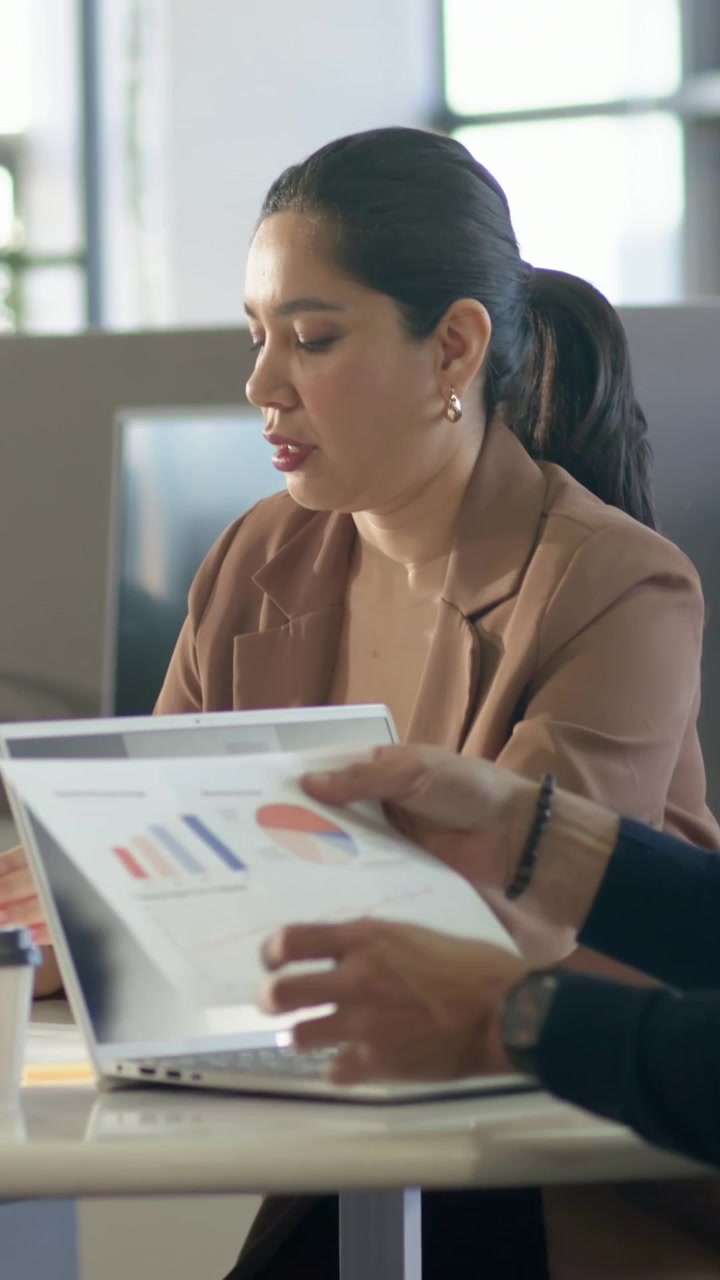 Vertical video: Receiving papers, woman in blazer scanning charts on laptop at meeting table