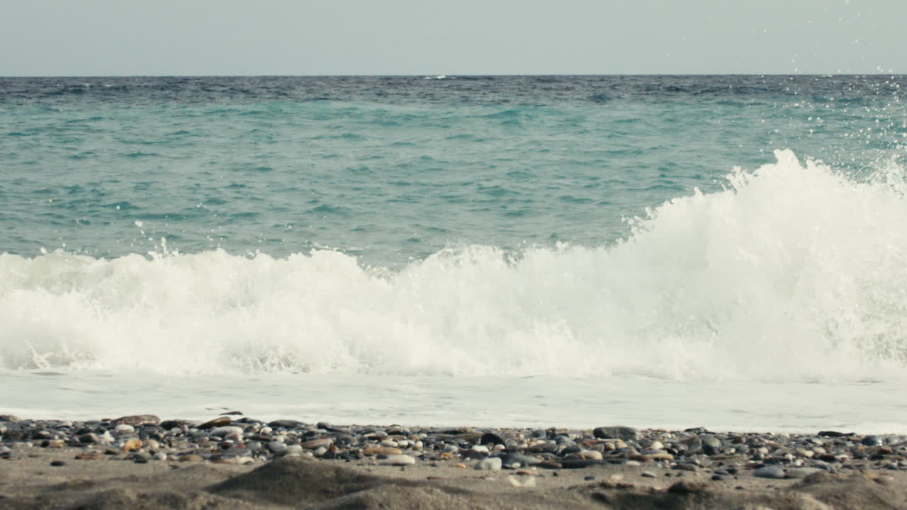 Low-angle slow motion shot of a clean wave chrashing on a pebbled shoreline, capturing natural rhythm and quiet anticipation in clear daylight