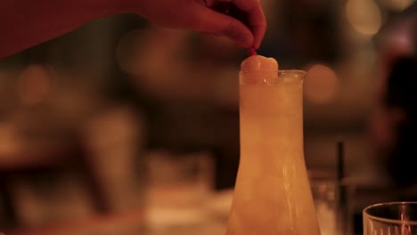a hand places a fruit garnish on a cocktail in a warm, ambient bar setting with soft lighting
