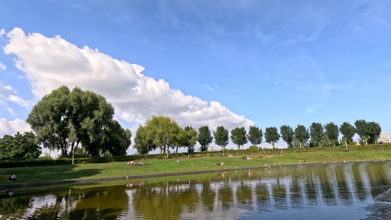 Wide shot of green park, tree line, blue sky, and pond reflections under daylight