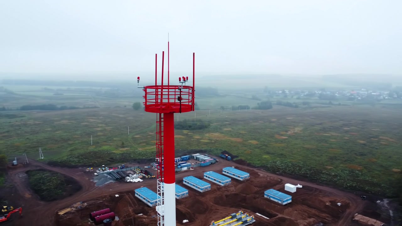 Alto traliccio rosso metallico per comunicazioni che si erge dal bordo di un'area di costruzione rurale, visto dall'alto con campi aperti, alberi ed edifici bassi che sfumano all'orizzonte.
