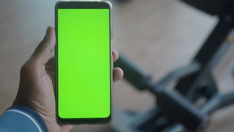 la mano de una persona sosteniendo un teléfono inteligente con una pantalla verde frente a un gimnasio