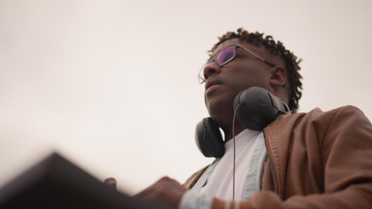 Uomo afroamericano vicino alla console, inquadratura cinematografica dal basso e focalizzata, cuffie appoggiate sull'attrezzatura, silhouette in primo piano, giacca di pelle, occhiali, atteggiamento serio da produttore, atmosfera urbana ambientale, cielo drammatico.