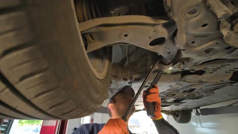 joven mecánico de automóviles en una estación de servicio de reparación inspeccionando la rueda del automóvil y el detalle de la suspensión de un automóvil levantado. vista de abajo.