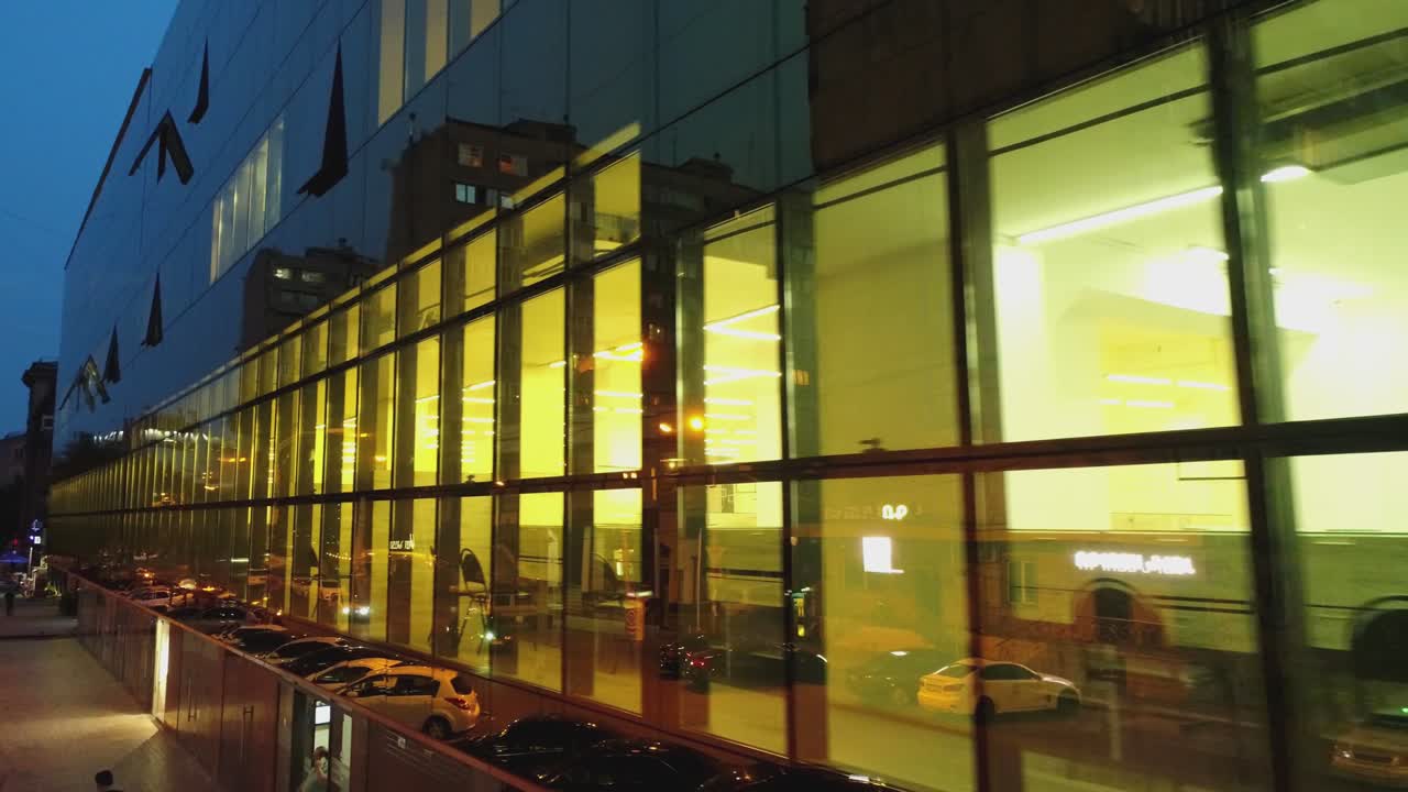 비즈니스 디스트릭트 건물의 4k 외관, 저녁이나 밤에 벽 거울 창문이있는 쇼핑몰, 거울 벽에 도시, 사람, 자동차의 반사, 드론 카메라 이동, 내부에서 일하는 사람들