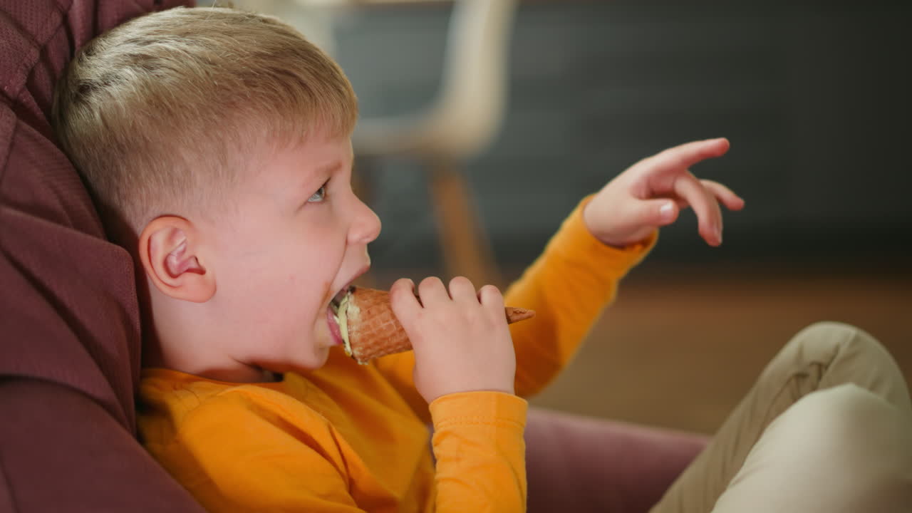 Side view close up of focused young boy in orange shirt licking ice cream cone while sitting comfortably indoors, with soft blurred background