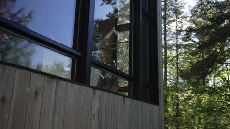 vista desde el exterior a través de la ventana de cristal de un hombre que se relaja en una hamaca dentro de la casa junto al bosque