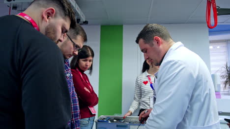 man in white robe shows the spine dummy and tool for surgery. students stand around listening to educator talking about neurosurgery.
