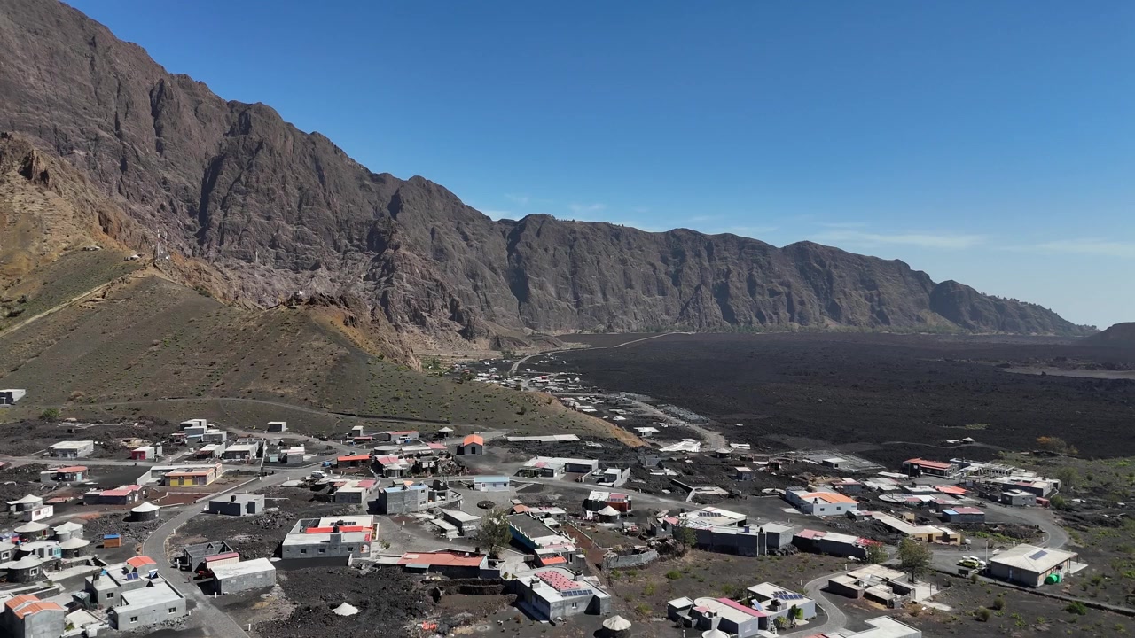 Perspectiva aérea do assentamento de Chã das Caldeiras na base da montanha Bordeira, com casas locais construídas no terreno vulcânico escuro e campos de lava da Ilha do Fogo, Cabo Verde.