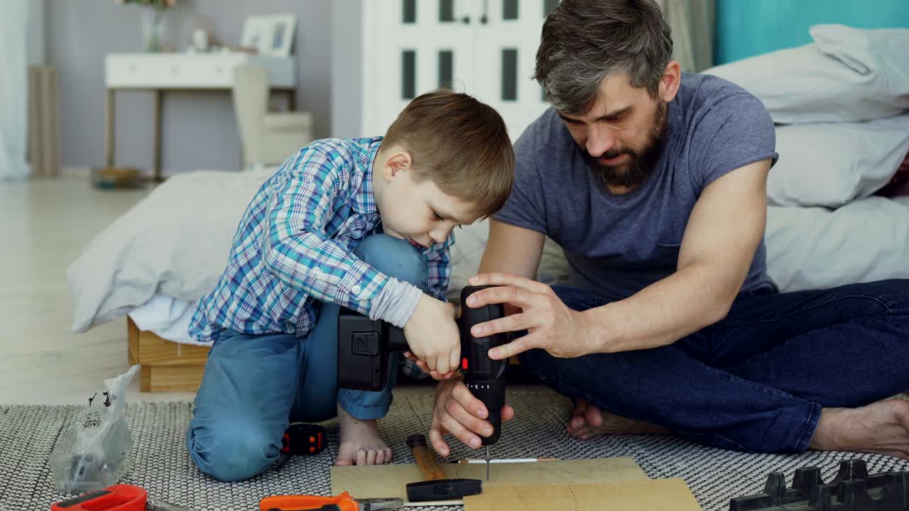Careful father is teaching his son to work with electric screwdriver while son is trying to use screw gun and fix screw in pieces of wood. Construction and family concept.