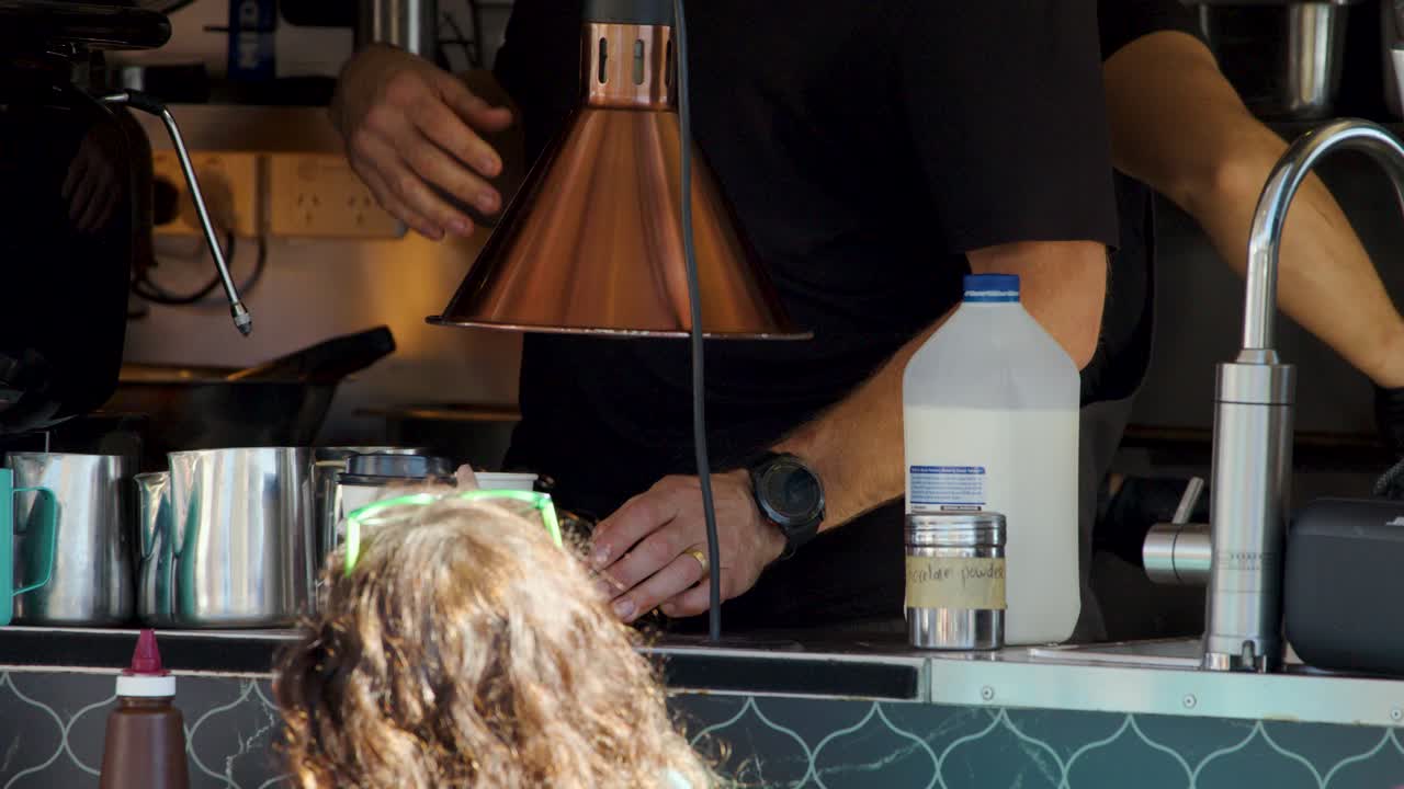 Barista pours milk into coffee cup at sunlit cafe counter, observed by customer, steady camera
